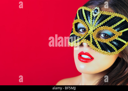 Hermosa morenita mujer vistiendo una máscara masquerade negro y dorado y el lápiz labial rojo brillante contra un fondo de color rojo. Foto de stock