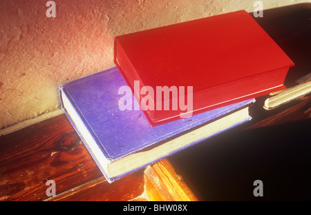 Uno rojo y uno azul libro sentado en la repisa de madera en las cálidas noches de luz solar y que generan largas sombras en crema de textura pared Foto de stock