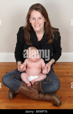 Retrato de una joven feliz Madre sentada sobre un piso de madera sosteniendo un recién nacido sano baby boy