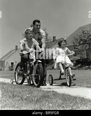 1950 padre empujando BOY EN BICICLETA CON RUEDAS DE ENTRENAMIENTO Y LA CHICA EN EL BARRIO TRICICLO ACERA Foto de stock