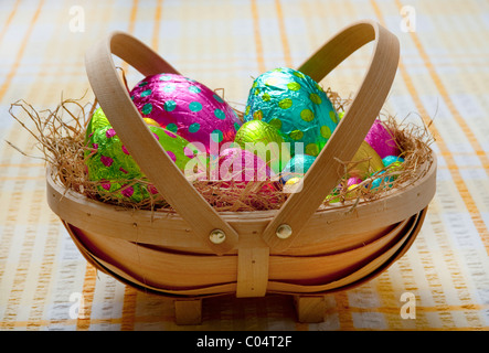 Pequeña cesta llena de madera tradicionales con heno y coloridos huevos de pascua de chocolate envuelto en un mantel blanco y amarillo Foto de stock