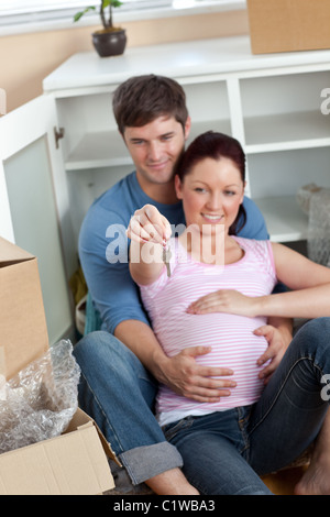 Encantado par esperando un bebé sentado en el suelo y mantenga la llave de su nueva casa Foto de stock