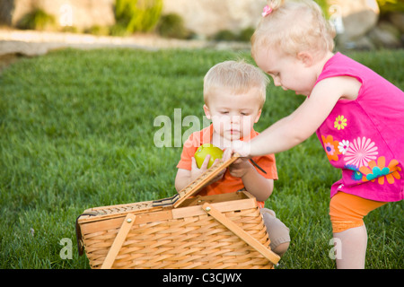 Hermano y hermana gemela lindos bebés jugando con Apple y cesta de picnic en el parque.
