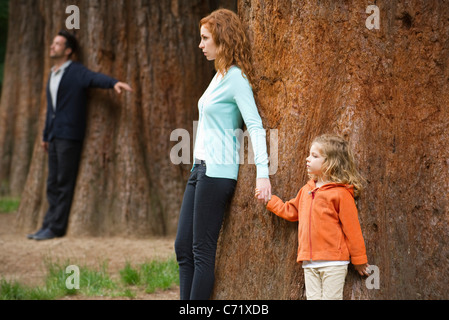 Madre e hija RECOSTADA contra el árbol, padre separado permanente en segundo plano. Foto de stock