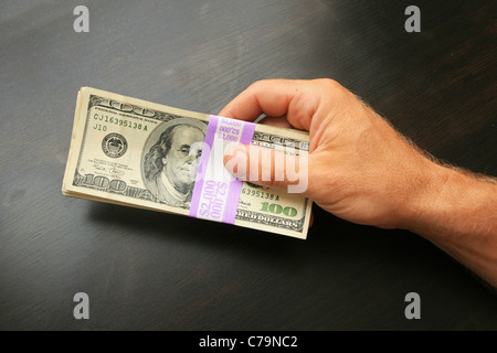Un mans mano sostiene dos mil dólares en billetes de cien dólares sobre una mesa de madera oscura Foto de stock