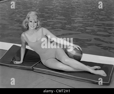 1960 rubia sonriente mujer vistiendo el traje de baño en piscina posar la mano sobre la pelota de playa