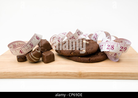 Galletas y chocolates en una tabla de cortar con medida representando el concepto que comer galletas y dulces se amontonan en las pulgadas Foto de stock