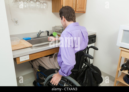 Hombre en silla de ruedas con lesión de la médula espinal preparando para lavar el vidrio en un fregadero accesible Foto de stock