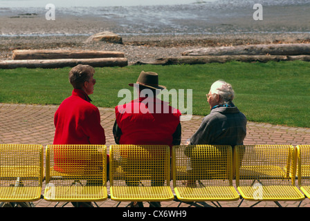 Tres Personas Mayores / amigos sentado en un banco y hablar, a lo largo de un paseo marítimo pasarela y Waterfront Park Foto de stock