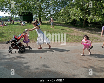 Una mujer caminando empujando un cochecito con su joven hija caminando por detrás en una ruta en el parque de Hampstead Heath, Londres NW3 UK KATHY DEWITT Foto de stock