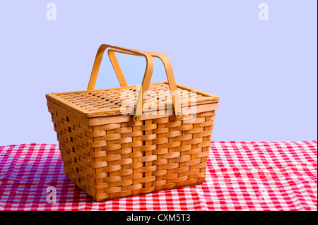 Una cesta de picnic de antigüedades de mimbre marrón sobre un mantel de tela de algodón a cuadros delante de un cielo azul Foto de stock