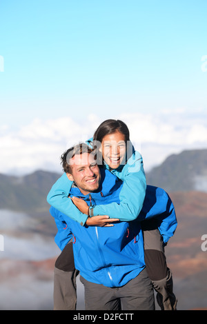 Alegre sonriendo interracial senderismo pareja disfrutando piggyback paseo en alta montaña Foto de stock