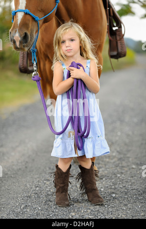 Poco chica rubia posando con su gran caballo sobre un carril del país, seguros de posar con mirada constante, Sol Azul Vestido y botas altas. Foto de stock