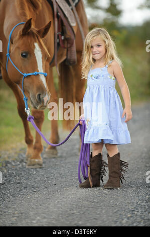 Poco chica rubia llevando su caballo grande abajo de un carril del país, sonriente y feliz tres años vistiendo vestido azul y western bo Foto de stock