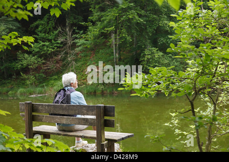 Mujer mayor walker descansando sentado en un banco bajo la lluvia en un lugar tranquilo en un entorno tranquilo del lago Taubensee Ruhpolding Chiemgau Baviera Alemania Foto de stock