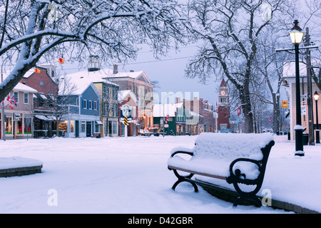 Canadá, Ontario, Niagara-on-the-Lake, Queen Street, temprano en la mañana de invierno, banco del parque cubierto de nieve que muestra una calle principal con tiendas. Foto de stock