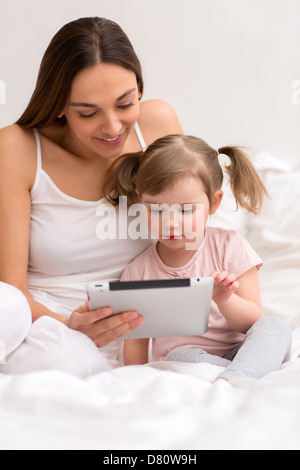 Niña jugando tablet con su mamá en el dormitorio blanco