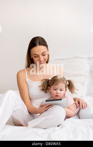 Niña jugando tableta digital con su mamá en el dormitorio blanco