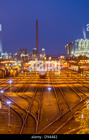 Estación de ferrocarril de la industria con iluminación naranja y azul cielo nocturno, una planta química en el fondo. Foto de stock