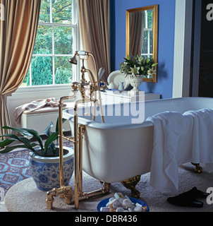 Grifos de latón autoestable y roll-top bañera en el cuarto de baño del país azul con crema de las cortinas en la ventana Foto de stock