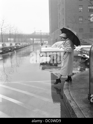 1950 MUJER EN CHUBASQUERO BOTAS SOMBRERO sosteniendo paraguas cruzando las calles de la CIUDAD EN MAL TIEMPO HÚMEDO Foto de stock