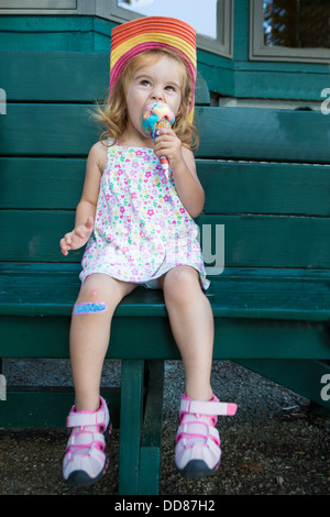 Poco lindo rubia en un sombrero de paja sentado en un banco de madera coloridas comiendo un helado en el sol de verano Foto de stock