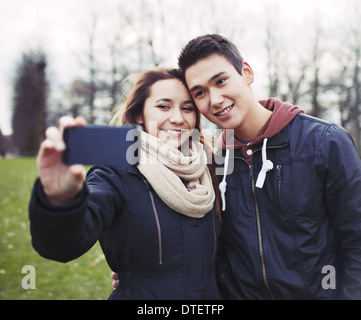 Linda pareja joven busca feliz mientras toma fotografías utilizando un teléfono inteligente en el parque. Mestizos adolescente y chica enamorada Foto de stock