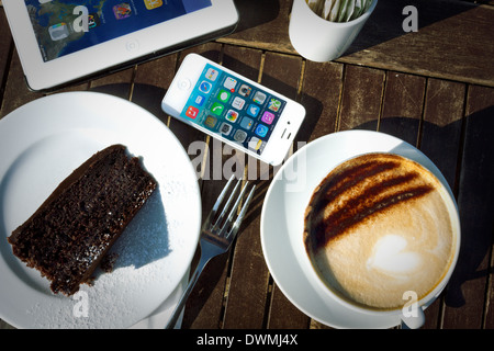 IPhone 4s, iPad 2 y torta y capuchino en la tabla en la luz del sol
