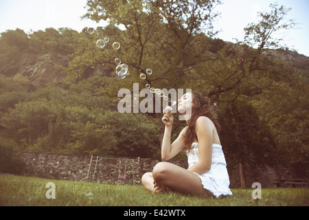 Mujer joven sentada piernas cruzadas en campo soplando burbujas