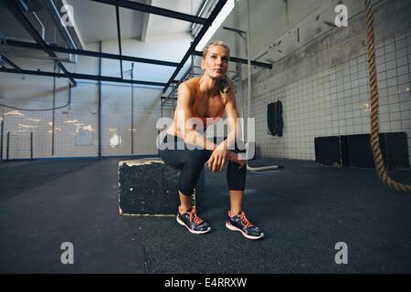 Mujer joven sentado en una caja en el gimnasio crossfit mirando a otro lado. Colocar joven atleta femenina del Cáucaso en el gimnasio.