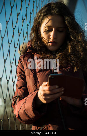 Close-up de adolescente de pie junto al eslabón de la cadena cerco, llevar abrigo y utilizando un teléfono inteligente, Alemania