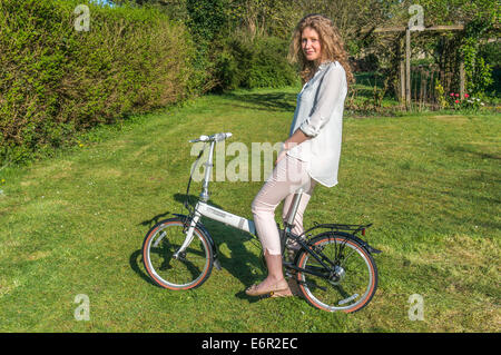 Una hermosa joven sentada sobre su bicicleta plegable Dahon en un jardín en Surrey, Inglaterra, Reino Unido.