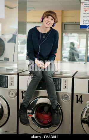 Mujer joven escuchando música en la parte superior de la lavadora, sonriendo Foto de stock