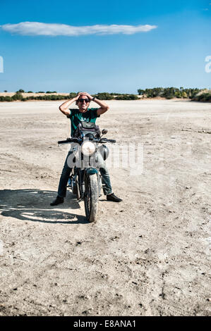 Mitad hombre adulto sentado en una motocicleta en la árida llanura, Cagliari, Cerdeña, Italia Foto de stock