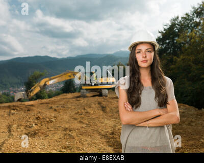 Retrato de joven mujer caucásica ingeniero en sitio de construcción con la excavadora en segundo plano.