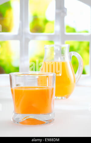 Saludable zumo de naranja recién exprimido en un vaso de vidrio con una jarra de cristal llena de jugo de pie delante de una ventana veraniego fresco Foto de stock