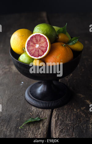 Pedestal tazón de fruta cítrica fresca surtida con la mitad de una sangre de color naranja aparece en la parte delantera, sobre un fondo de madera rústica con sombras Foto de stock