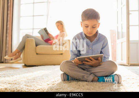 Boy utilizando digital tablet en piso con madre leyendo magazine en segundo plano.