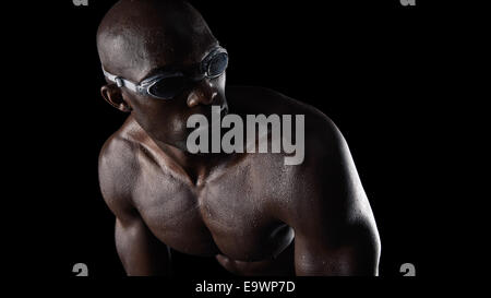 Foto de estudio de nadador masculino africano gafas protectoras mirando por encima del hombro, contra un fondo negro. Musculoso joven atleta.