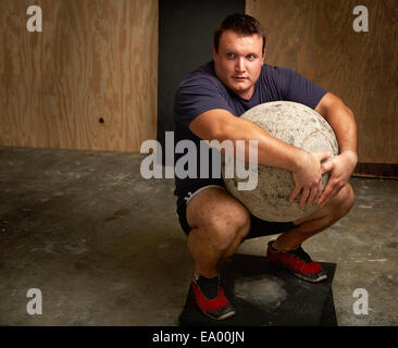 Retrato de macho joven levantador de pesas en cuclillas con atlas ball en el gimnasio Foto de stock