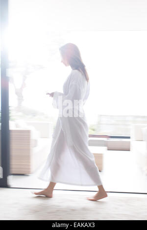 Mujer vistiendo albornoz blanco caminando por el corredor moderno con teléfono móvil Foto de stock