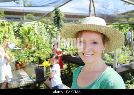 F Retrato mujer sonriente vistiendo sombrero en invernadero, niños en segundo plano.