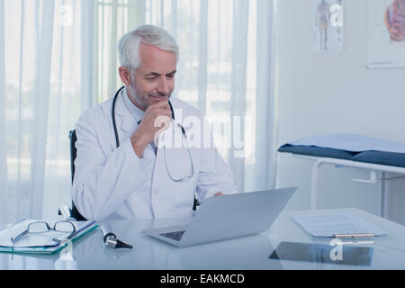 Sonriente doctor maduro sentado en una mesa con un portátil en la oficina