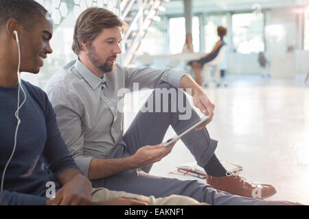 Dos hombres de negocios utilizando tablet digital y auriculares sentados en el piso en la oficina moderna