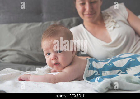 Retrato de pequeño bebé acostado en la cama con la madre sonriendo en segundo plano. Foto de stock