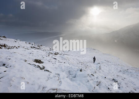 Un caminante solitario desciende de una montaña cubierta de nieve caminando hacia los rayos del sol que iluminan la nieve cayendo de nubes oscuras Foto de stock