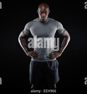 Retrato del joven muscular en ropa deportiva de pie con las manos en las caderas contra el fondo negro. Fuerte atleta africano. Foto de stock