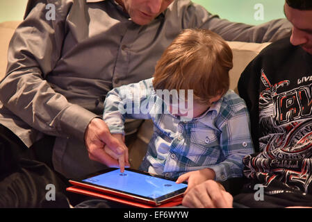 Un joven (2 1/2 años) jugando en un iPad sentado entre dos adultos que están ayudando a él jugar Foto de stock