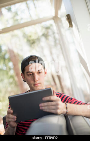 Hombre que llevaba una gorra de béisbol hacia atrás, sentado en un sofá, mirando a una tableta digital.