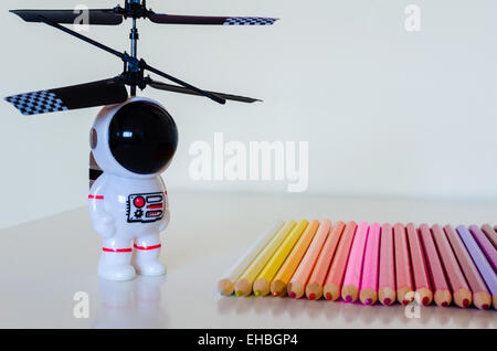 Un juguete infantil spaceman mira hacia un juego de lápices de colores sentado en una fila en una superficie blanca. Hay algunos reflejan los detalles.